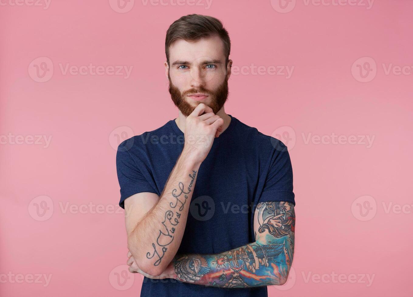 ung tänkande attraktiv rödskäggig kille med blå ögon, bär en blå t-shirt, innehar hans hand på hans haka och utseende eftertänksamt in i de kamera isolerat över rosa bakgrund. foto