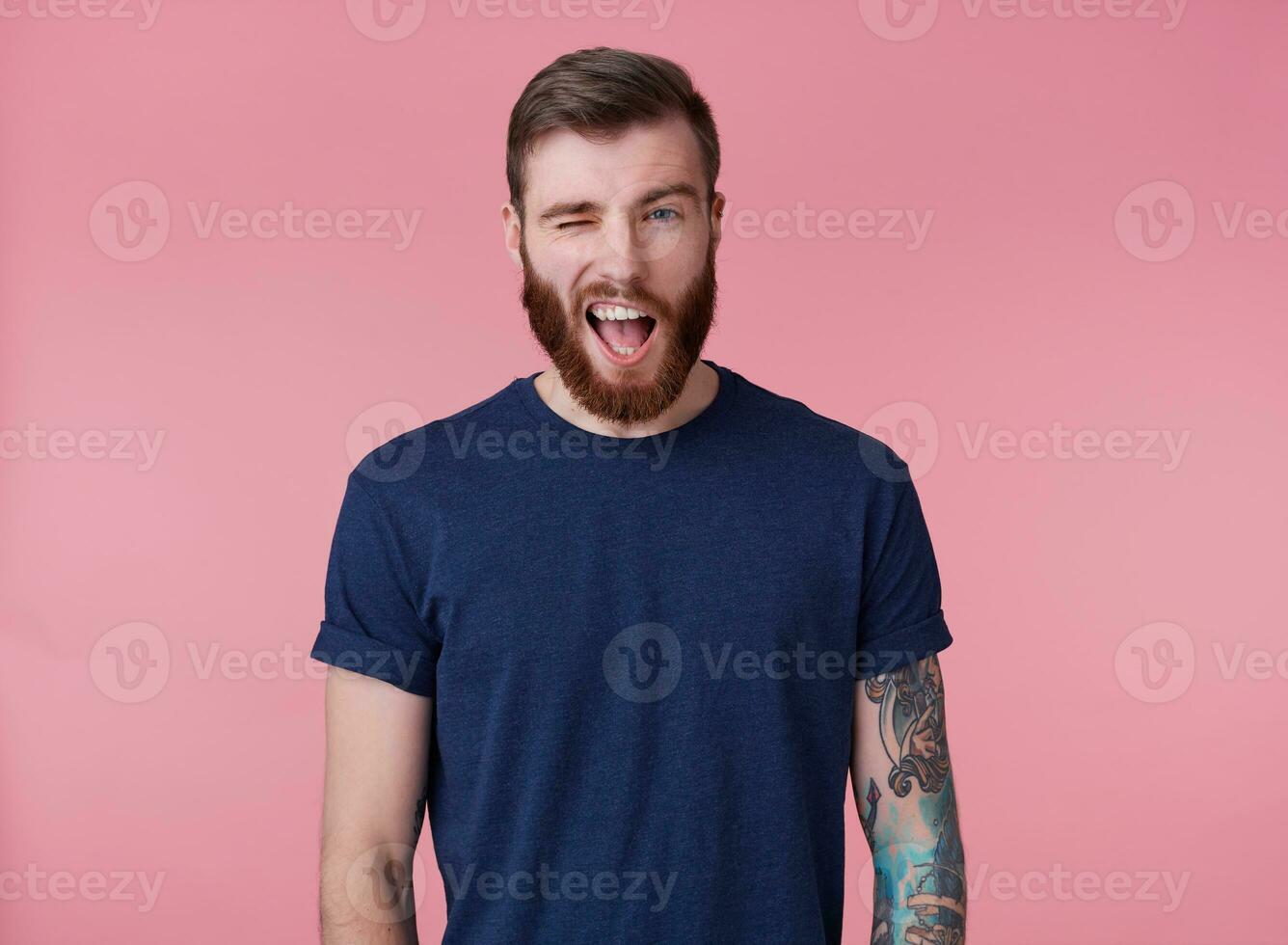 ung attraktiv rödskäggig kille med blå ögon, bär en blå t-shirt, ser på de kamera och blinkar, se Häftigt isolerat över rosa bakgrund. foto