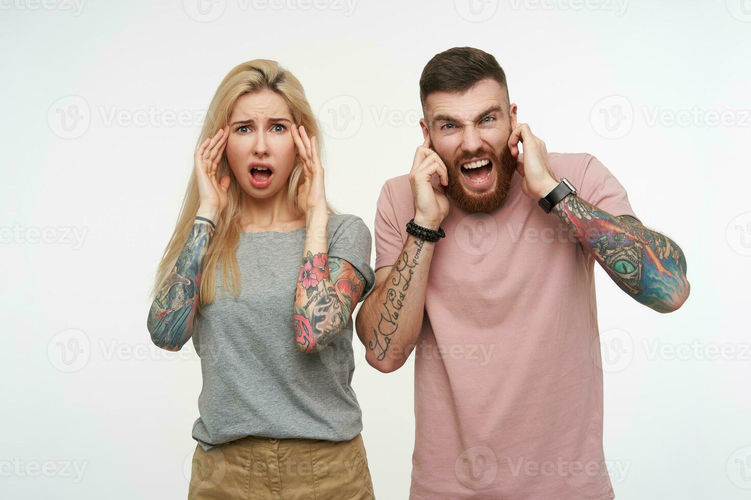 förvirrad ung blåögd Söt par i tillfällig t-tröjor höjning händer till deras ansikten och rynkar ögonbryn medan ser förvånad på kamera, isolerat över vit bakgrund foto