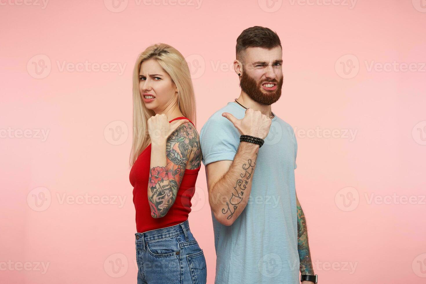 missnöjd ung Söt tatuerade människor som visar på varje Övrig med Uppfostrad händer och rynkar missnöjt ansikten medan ser på kamera, isolerat över rosa bakgrund foto
