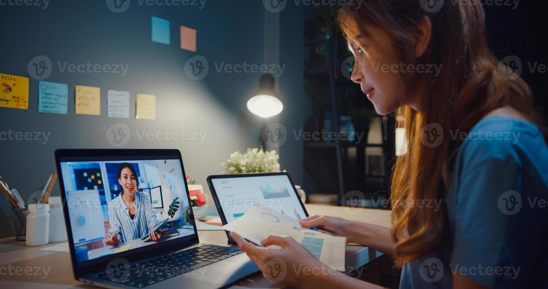 asiatisk kvinna som använder bärbar dator prata med kollegor om arbete i videosamtal medan hon arbetar hemma i vardagsrummet på natten. självisolering, social distansering, karantän för förebyggande av coronavirus. foto