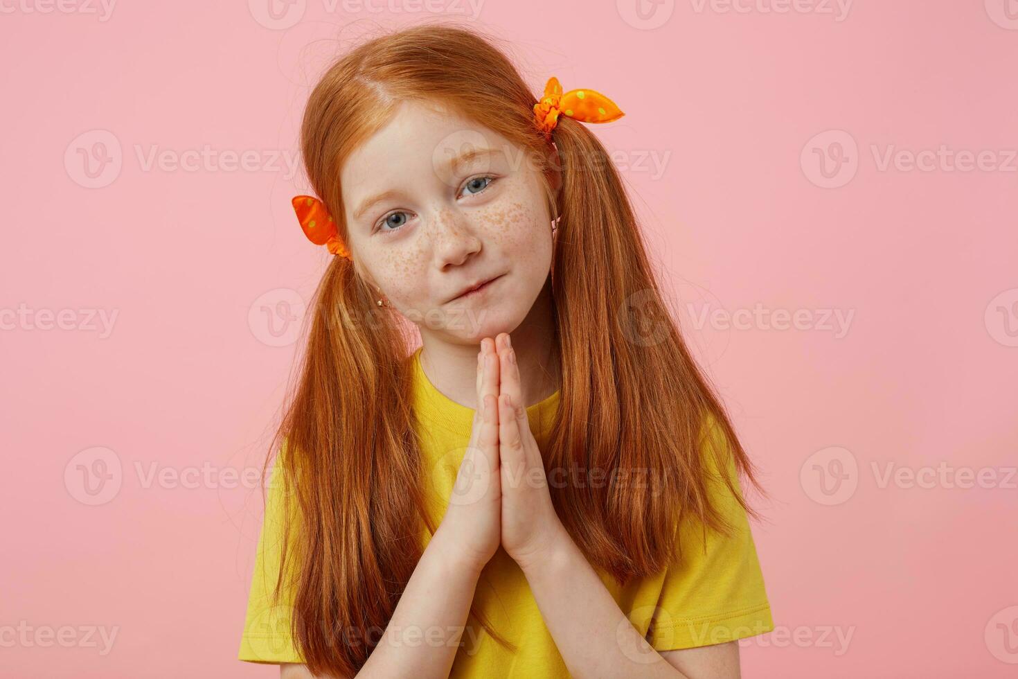 porträtt liten förlåt fräknar rödhårig flicka med två svansar, utseende in i de kamera och kupad händer tillsammans, byte gest, bär i gul t-shirt, står över rosa bakgrund. foto