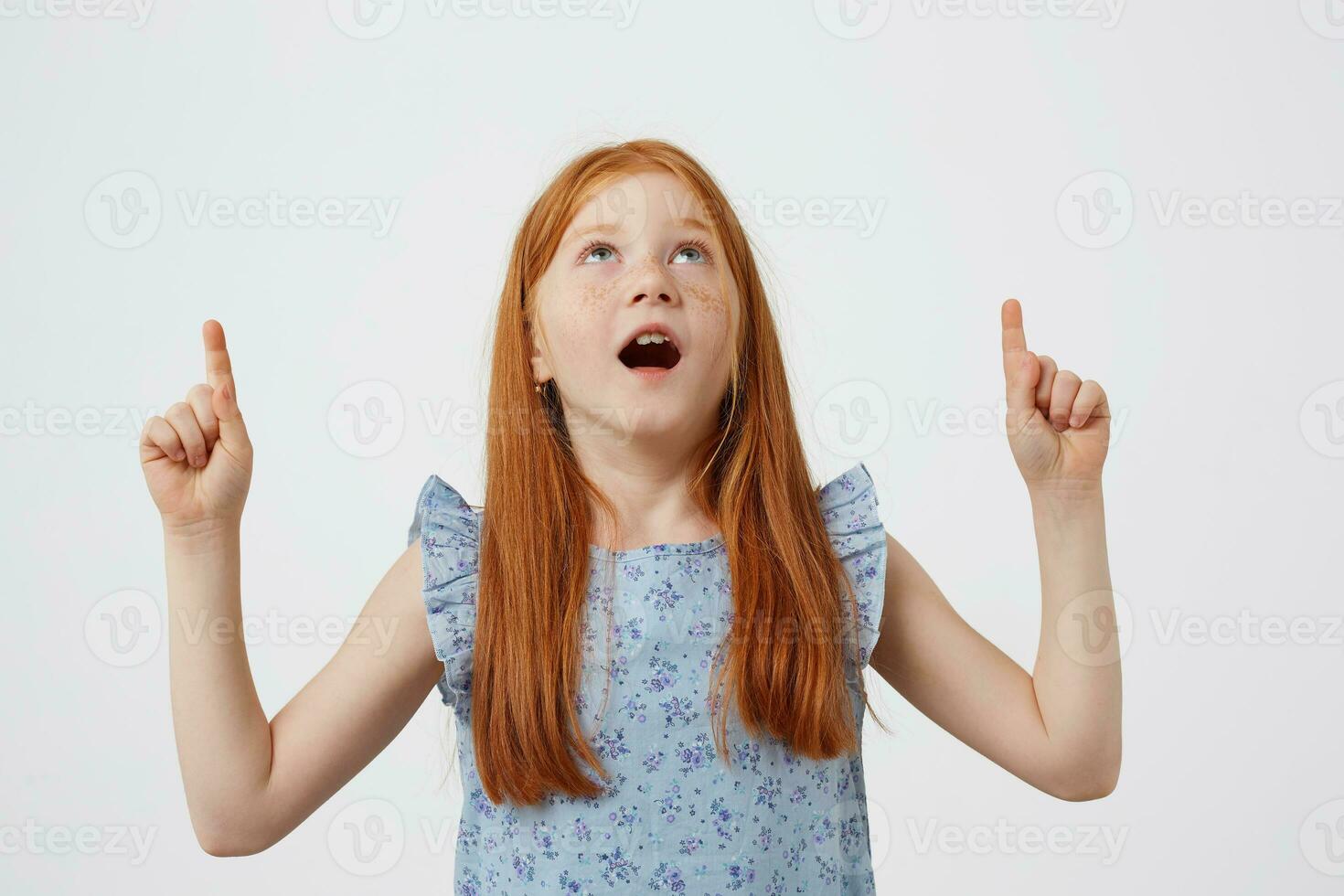 Foto av överraskad liten fräknar rödhårig flicka med två svansar, utseende upp och pekande fingrar på de kopia Plats, bred öppen mun och ögon, bär i blå klänning, står över vit bakgrund.