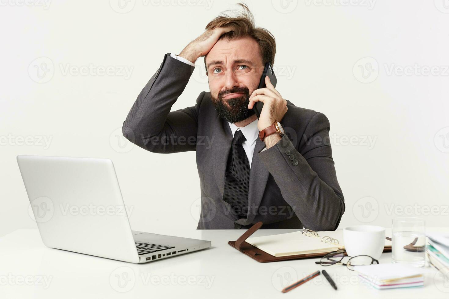 betonade skäggig brunett manlig Sammanträde på arbetssätt tabell och har spänd telefon konversation, rumling hans hår med förvirrad ansikte och ser förbryllad åt sidan, klädd i grå kostym foto