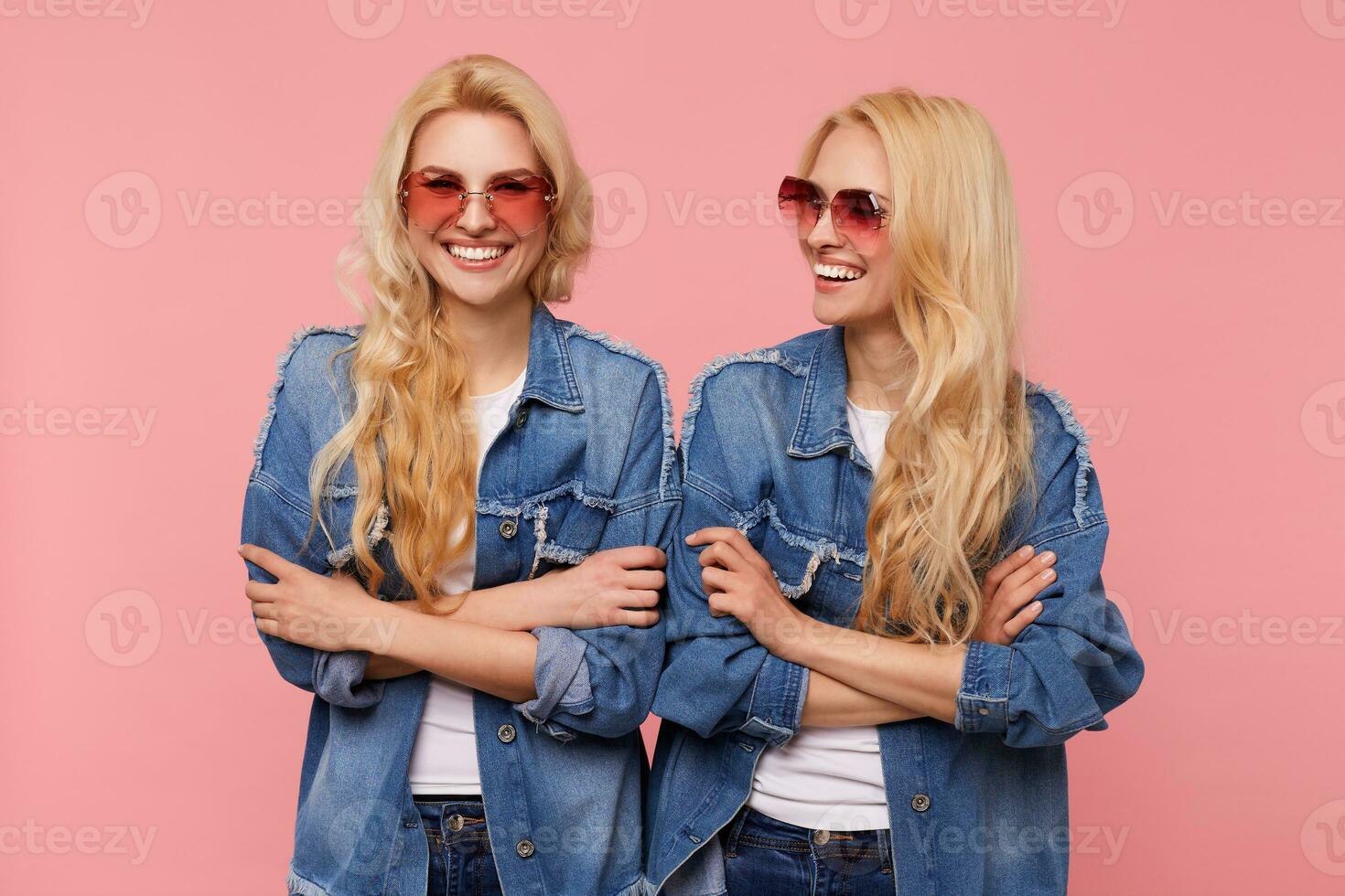 Lycklig ung lång håriga blond honor klädd i solglasögon och tillfällig kläder förvaring händer vikta och skrattande glatt medan Framställ över rosa bakgrund foto