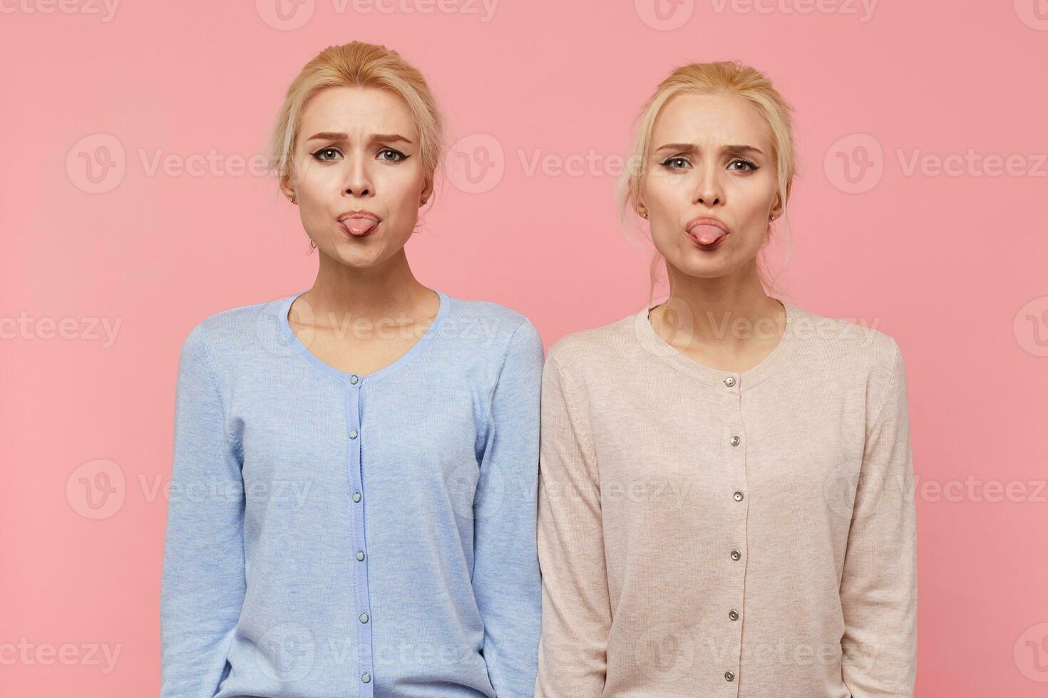 porträtt av skön ung blond tvillingar göra ansikten och visa tungor, ser på de kamera isolerat över rosa bakgrund. foto