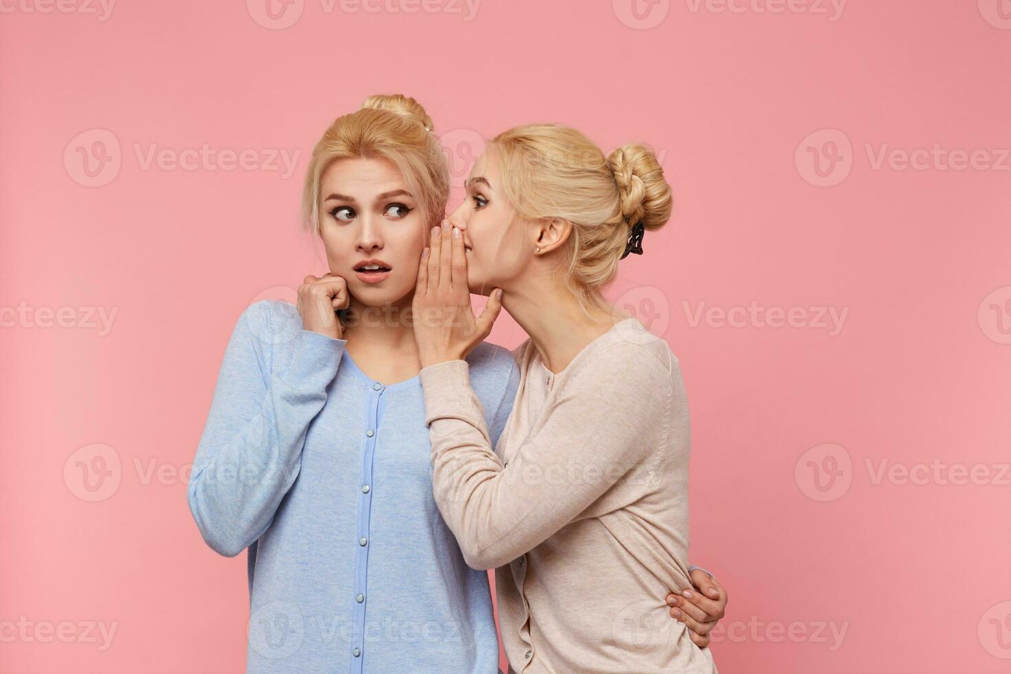 porträtt av söt tvillingar blondiner, de flicka berättar henne syster de otrolig Nyheter handla om de galen rabatter, de andra ett öppnad henne mun i överraskning, står över rosa bakgrund. foto