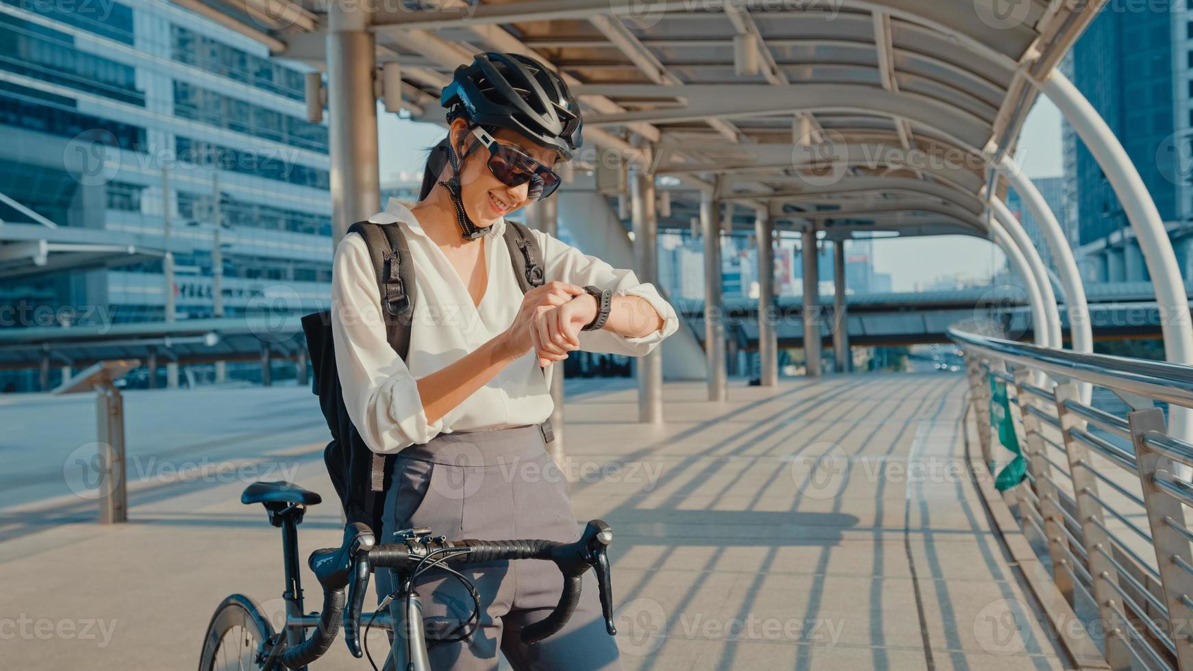asiatisk affärskvinna med ryggsäck cykel le smartwatch i stadsgatan gå till jobbet på kontoret. sportflicka använder sin klockapp för fitnessspårning. pendla till jobbet, affärspendlare i staden. foto