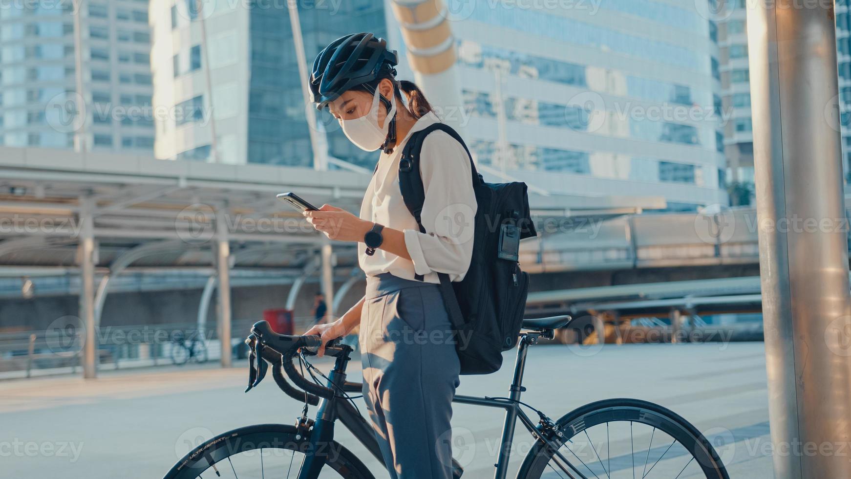 asiatisk affärskvinna bär ryggsäck bär antivirusskyddsmask ta cykelpromenad och kolla telefonen i stadsgatan gå till jobbet på kontoret. pendla till jobbet, affärspendlare för covid-19-konceptet. foto