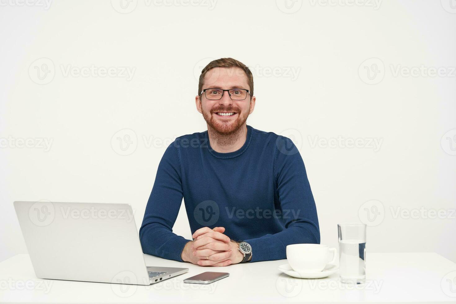 glad ung Söt skäggig ljushårig manlig i glasögon förvaring vikta händer på tabell medan ser lyckligt på kamera med charmig leende, isolerat över vit bakgrund foto