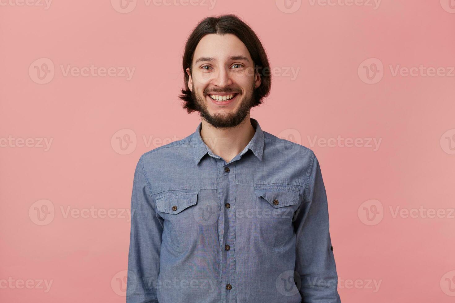 Foto av leende skäggig ung man med lång kammad mörk hår i denim skjorta, ser på de kamera isolerat över rosa bakgrund.