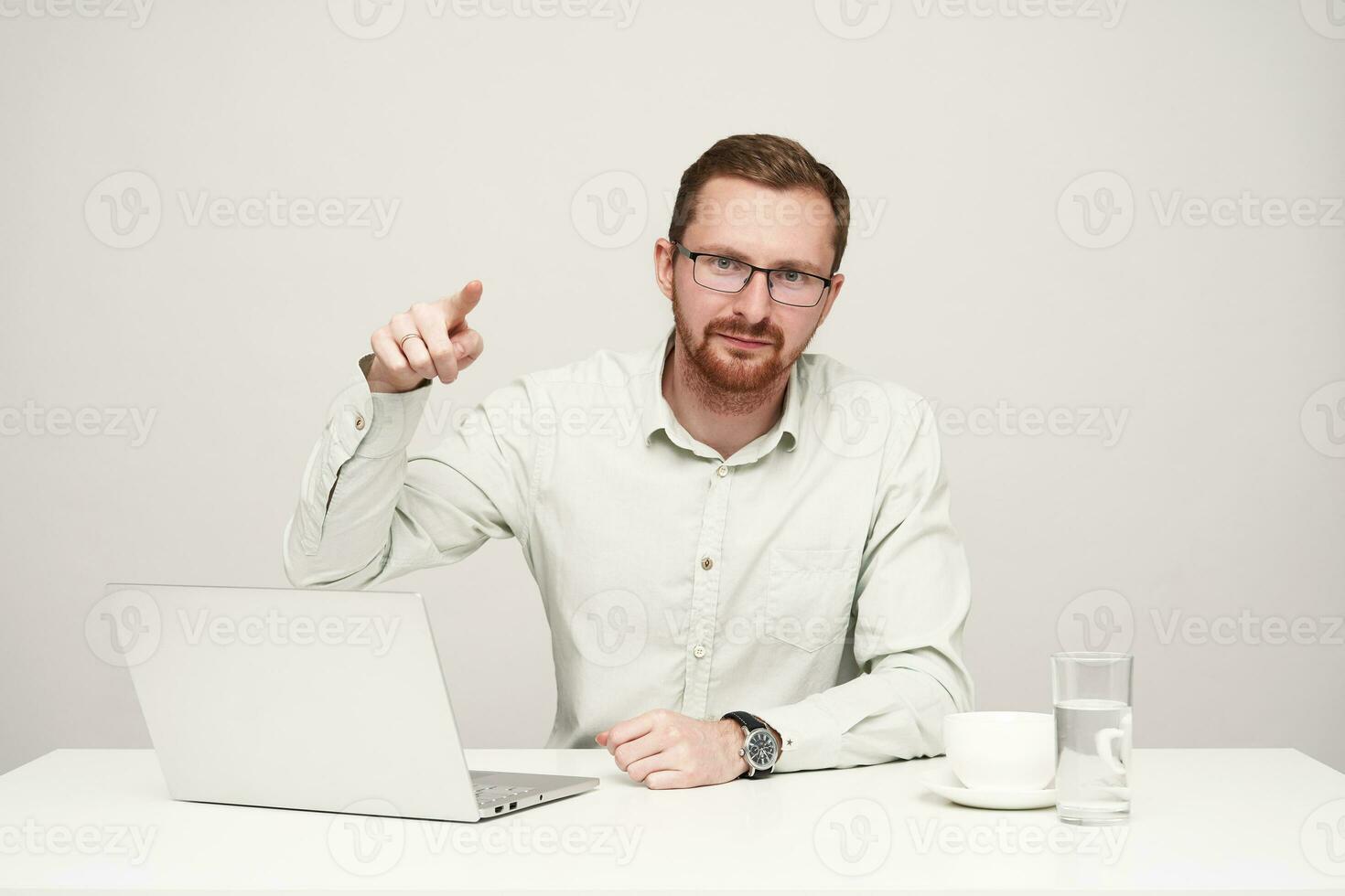 horisontell skott av ung skäggig korthårig manlig i glasögon förvaring hand Uppfostrad medan pekande på kamera med pekfinger, Sammanträde över vit bakgrund foto