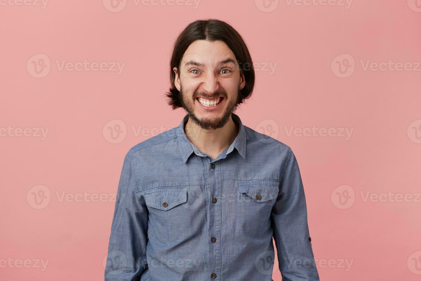 Foto av arg skäggig ung man med lång kammad mörk hår i en dålig humör, utseende arg och missnöjd isolerat över rosa bakgrund.