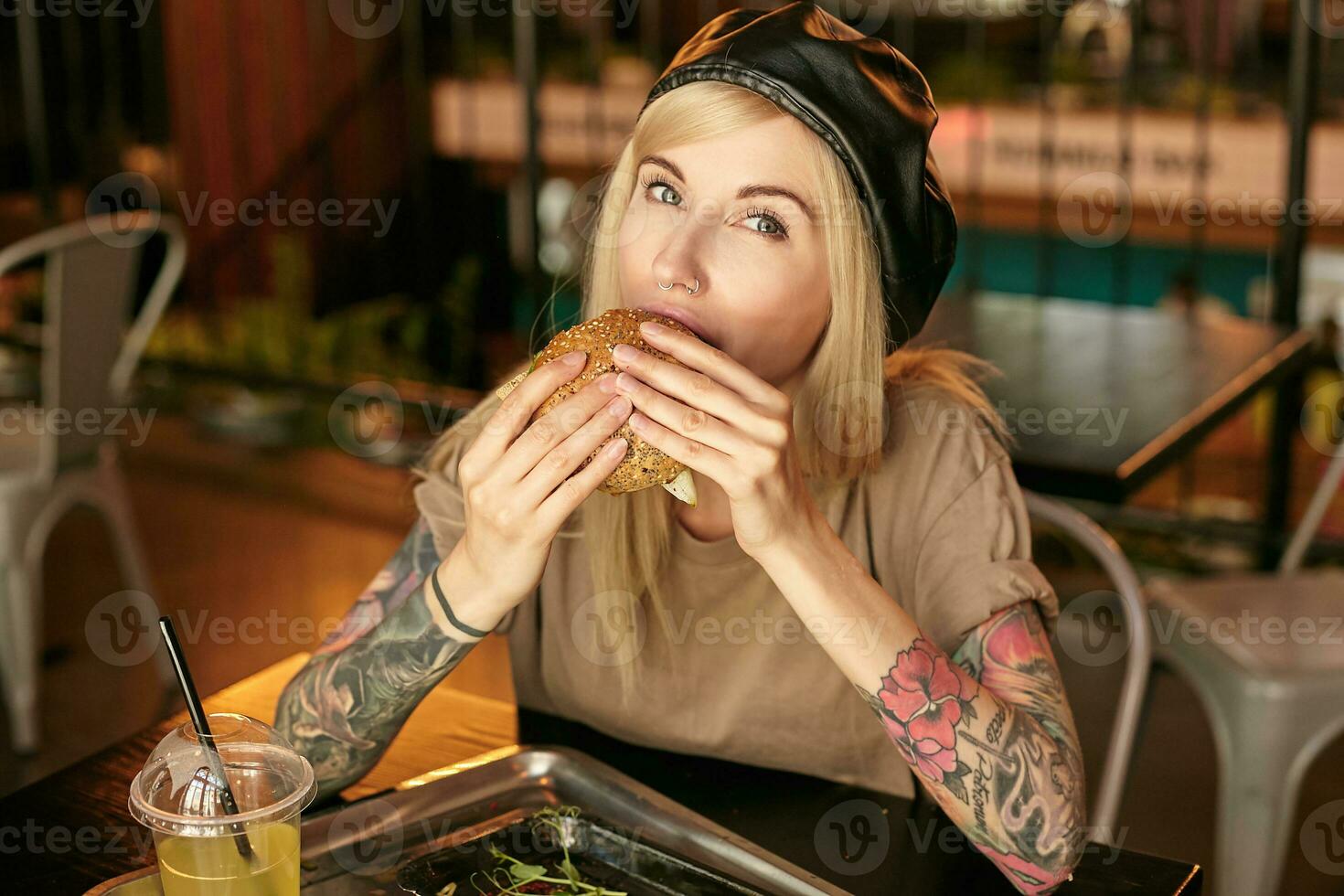 charmig tatuerade blond kvinna med lång hår ser på kamera medan påfrestande hamburgare i stad Kafé under lunch ha sönder, bär beige t-shirt och läder svart basker foto