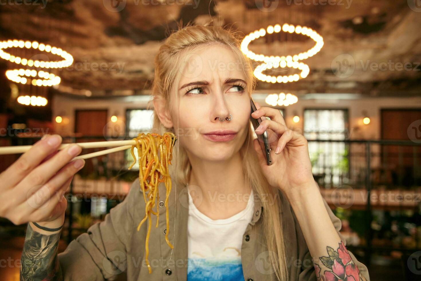 porträtt av allvarlig ser tatuerade blond kvinna med tillfällig frisyr innehav spaghetti med trä- pinnar medan talande på pphone, bär beige skjorta och vit t-shirt foto