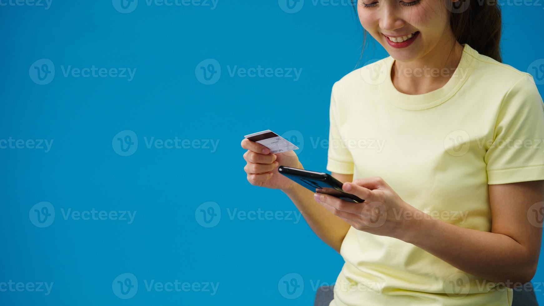 ung asiatisk dam som använder telefon och kreditbankkort med positivt uttryck, ler brett, klädd i vardagskläder och står isolerad på blå bakgrund. glad förtjusande glad kvinna jublar över framgång. foto