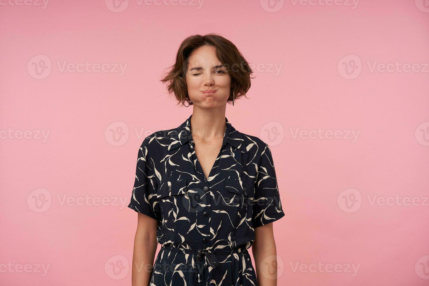 porträtt av rolig ung kvinna med kort brun hår puffande ut kinder och blinka på kamera, varelse i hög anda, stående över rosa bakgrund med händer ner foto
