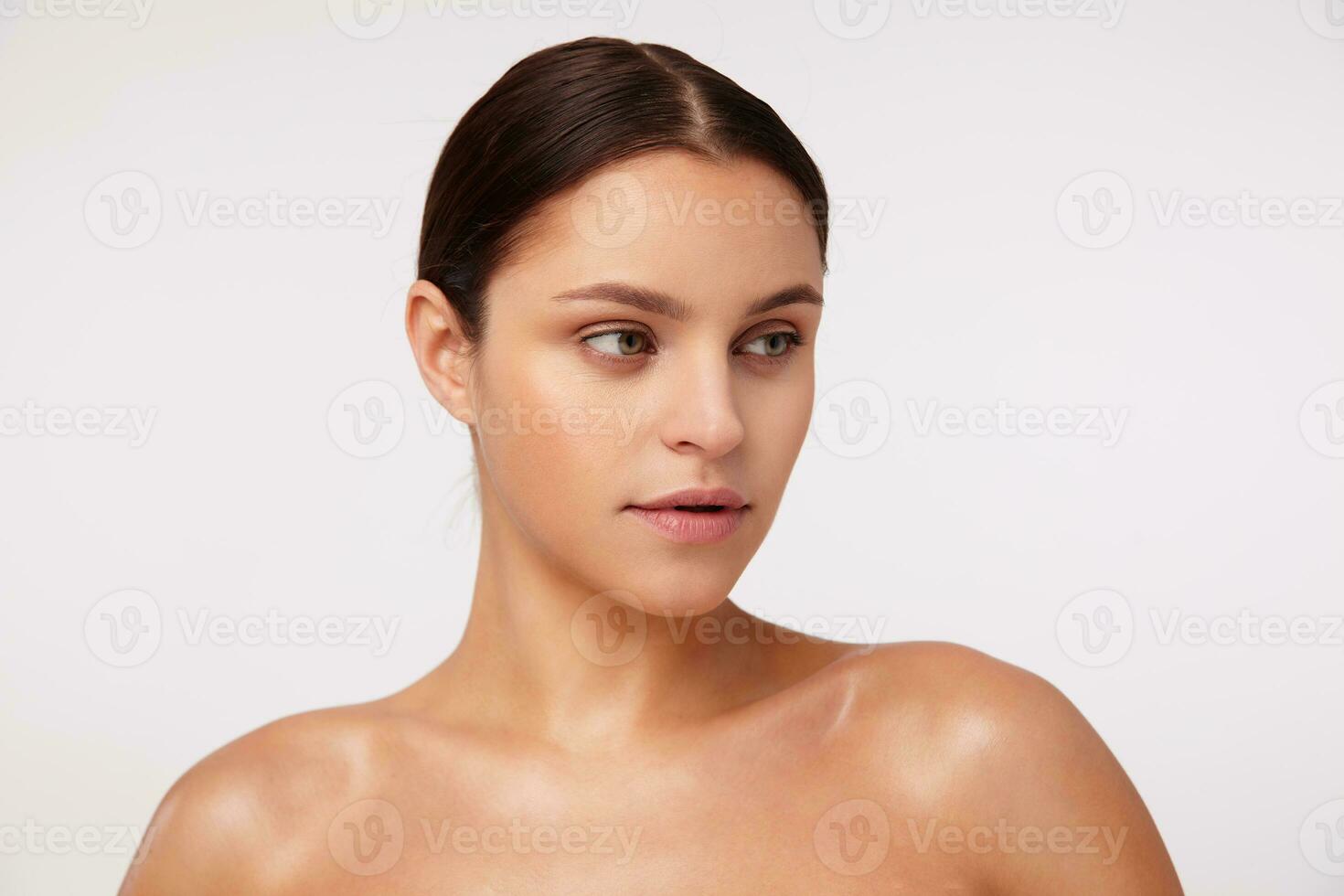 närbild av charmig ung brunett kvinna bär naturlig smink och tillfällig frisyr medan Framställ mot vit bakgrund, ser åt sidan med lugna ansikte foto