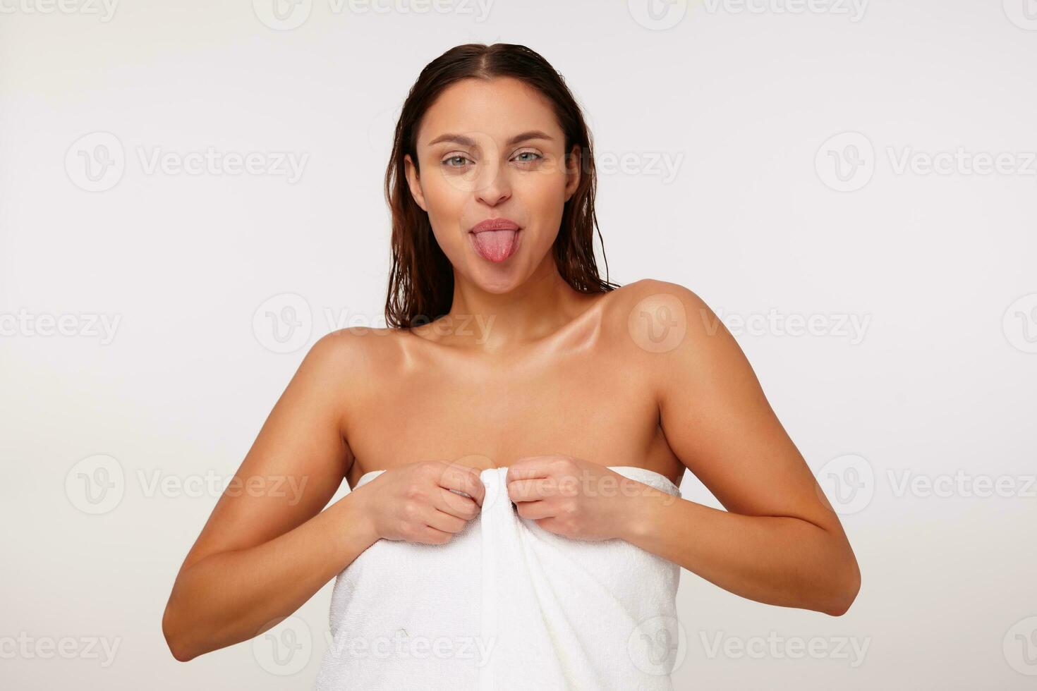 inomhus- Foto av ung skön glad mörk håriga kvinna som visar tunga medan ser positivt på kamera, stående över vit bakgrund i bad handduk