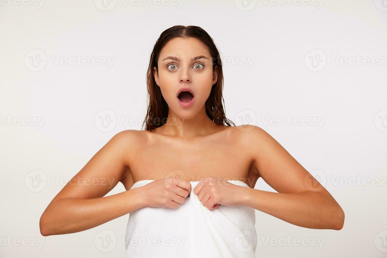 porträtt av förvånad ung Söt brunett kvinna med våt hår ser på kamera med bred ögon och mun öppnad medan Framställ över vit bakgrund efter dusch foto