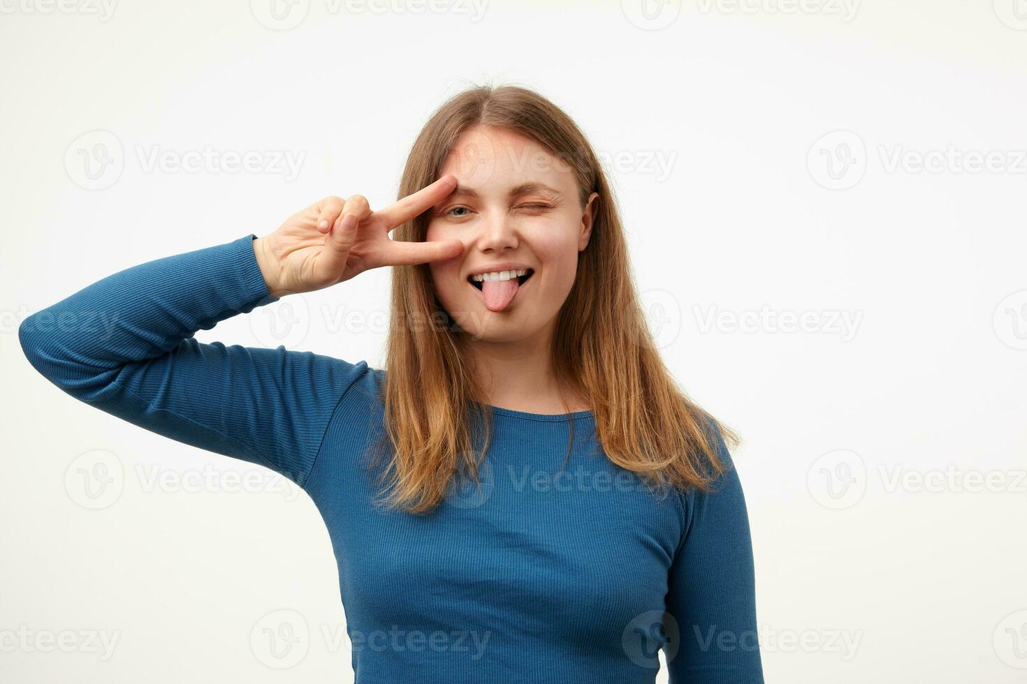 glad ung Söt lång håriga kvinna höjning hand med seger gest till henne ansikte och som visar glatt tunga på kamera medan stående över vit bakgrund foto