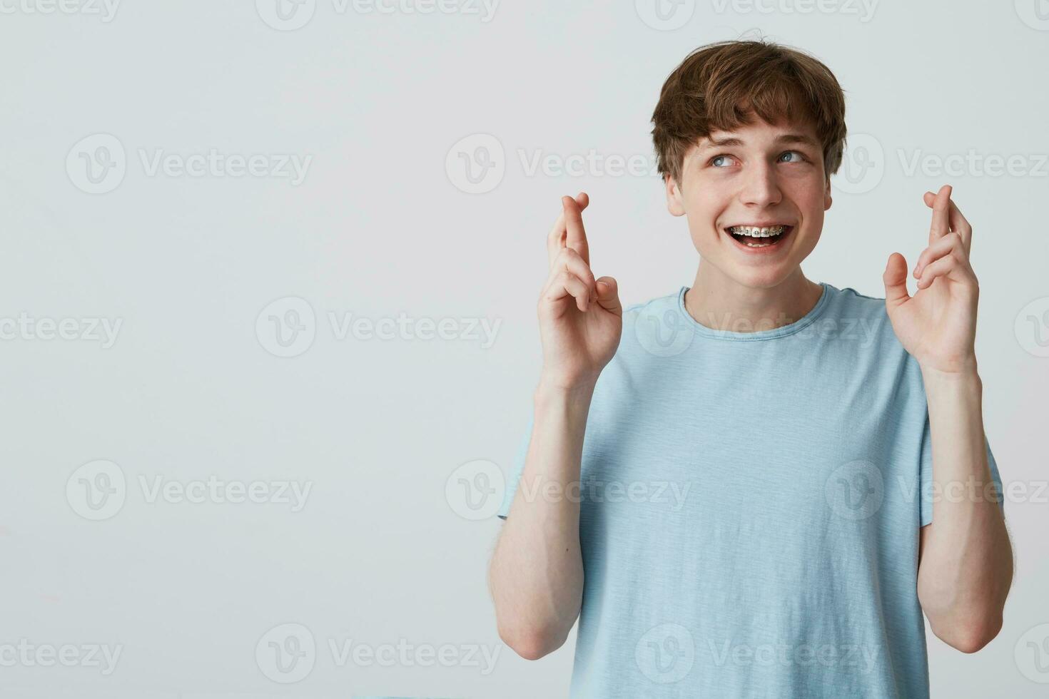 tonåring utseende bort, önskar något trevlig, händer upp och fingrar vriden, med tandställning på tänder, bär blå t-shirt, blank kopia Plats för din PR innehåll isolerat över vit bakgrund foto