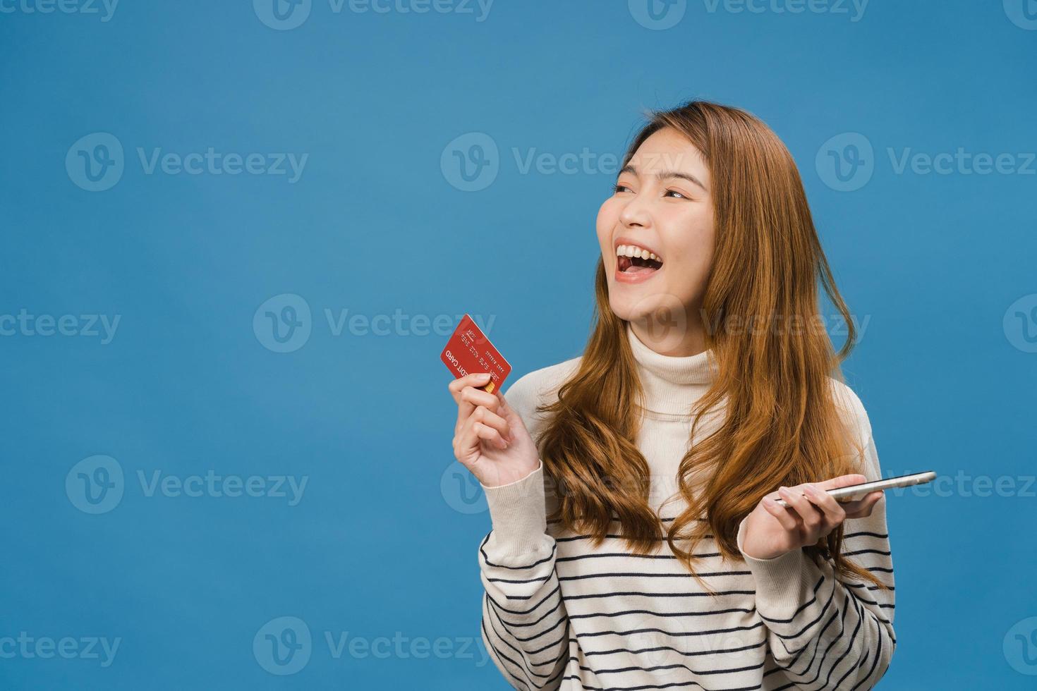 ung asiatisk dam som använder telefon och kreditbankkort med positivt uttryck, ler brett, klädd i vardagskläder och står isolerad på blå bakgrund. glad förtjusande glad kvinna jublar över framgång. foto