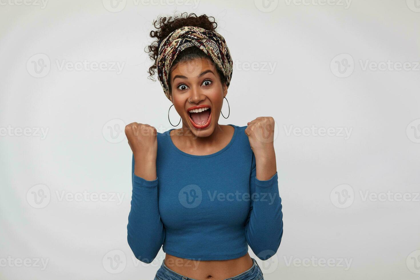 inomhus- Foto av ung upprörd mörk håriga lockigt kvinna med röd mun höjning lyckligt henne nävar medan fröjd handla om något, isolerat över vit bakgrund