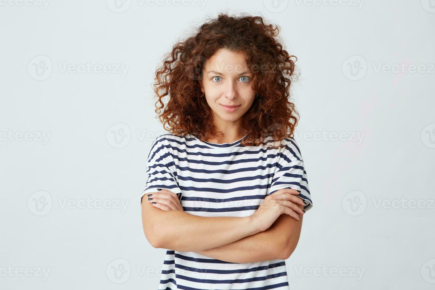 porträtt av Lycklig attraktiv ung kvinna med lockigt hår bär randig t skjorta utseende självsäker och står med vapen korsade isolerat över vit bakgrund foto