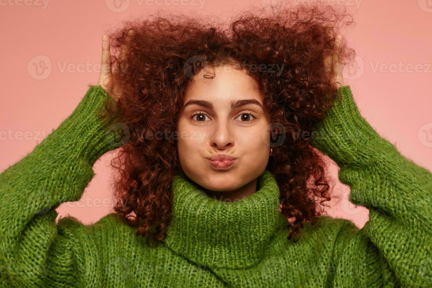 ung lady, Söt kvinna med ingefära lockigt hår. bär grön turtleneck Tröja och rörande henne hår, puffar ut kinder. tittar på på de kamera isolerat, närbild över pastell rosa bakgrund foto