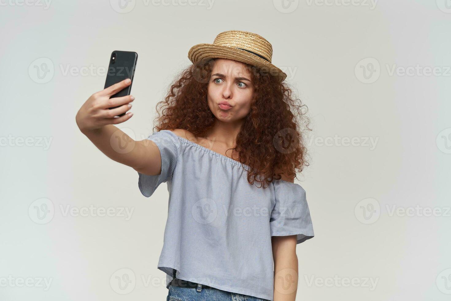 Tonårs flicka, Lycklig ser kvinna med lockigt ingefära hår. bär randig off-shoulders blus och hatt. tar en selfie på en smartphone och puta med läpparna mun. stå isolerat över vit bakgrund foto