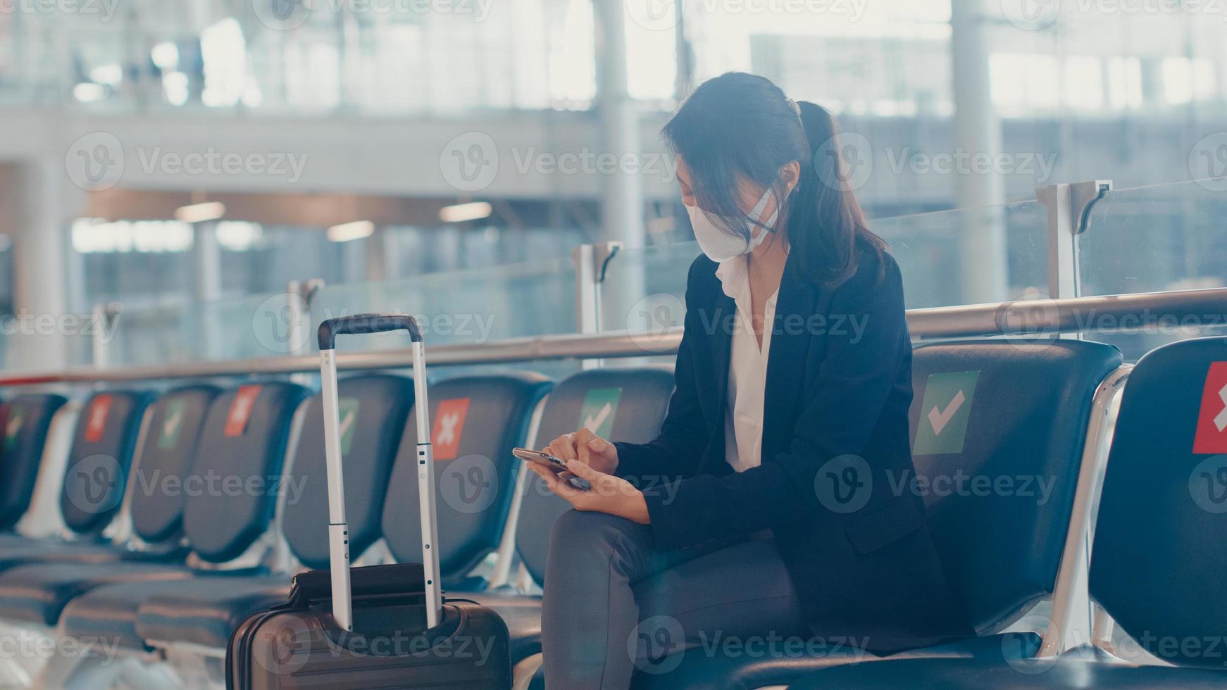 asiatisk affärs damresenär bär kostym sitter med resväska och använder smart telefon chattmeddelande i bänk vänta på flygning på flygplatsen. affärsresependlare i covid -pandemi, affärsresekoncept. foto