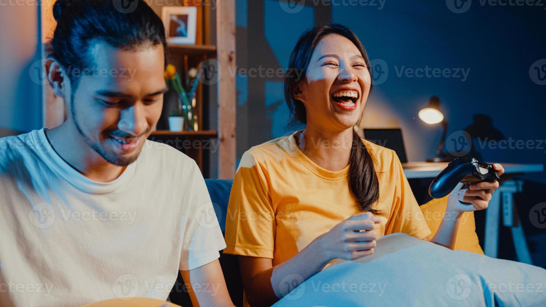 lyckligt asien ungt par man och kvinna sitter i soffan använda joystick -kontroller spela videospel tillbringa rolig tid tillsammans i vardagsrummet på natten. asiatiska gift par familj livsstil, stanna hemma koncept. foto