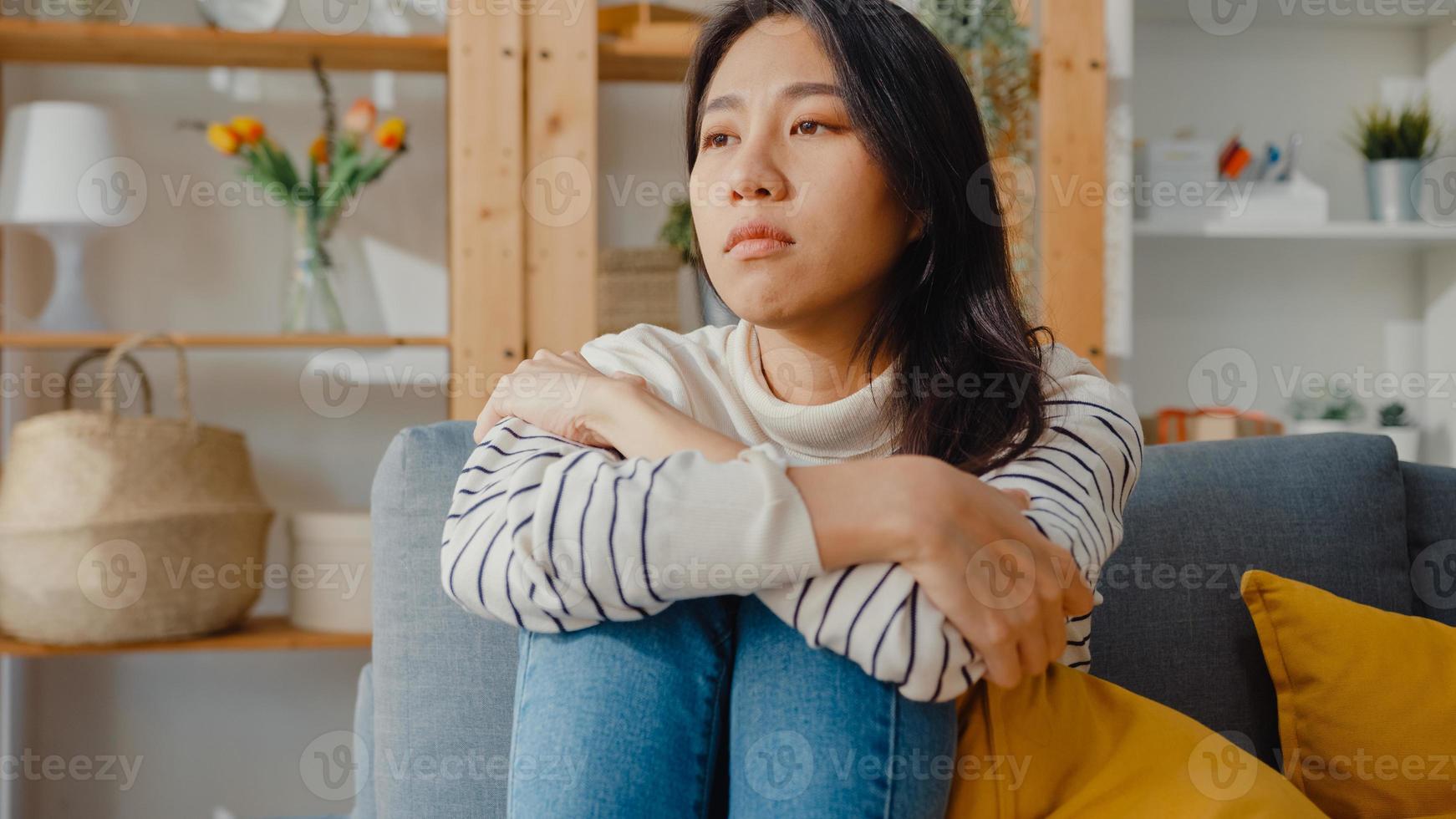 tankeväckande asiatisk dam sitter och omfamnar knän i soffan i vardagsrummet hemma och tittar ut utanför med ensam känsla, ledsen deprimerad tonåring tillbringar tid ensam stannar hemma, social distans, coronavirus -karantän. foto