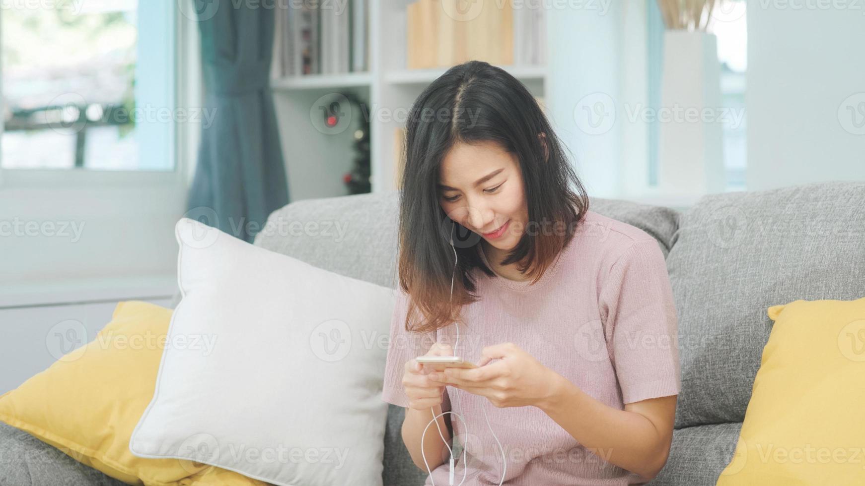 asiatisk kvinna som lyssnar musik och använder smart telefon, kvinnan använder slappningstid som ligger på hemmasoffan i vardagsrummet hemma. glad kvinnlig lyssnar musik med hörlurar koncept. foto