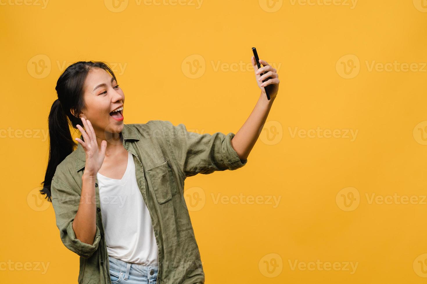 le bedårande asiatisk kvinna som gör selfie foto på smart telefon med positivt uttryck i vardagskläder och stativ isolerad på gul bakgrund. glad förtjusande glad kvinna jublar över framgång.