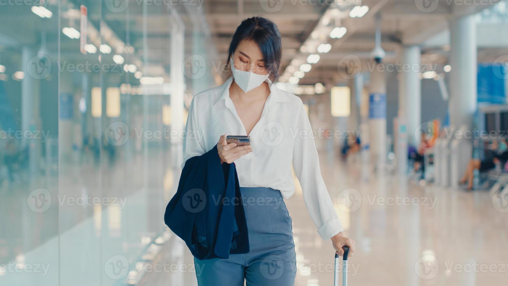 asiatisk affärsflicka använder smart telefon för att checka in boardingkort promenad med bagage till terminal på inrikesflyg på flygplatsen. affärspendlare covid -pandemi, affärsresor social distanseringskoncept. foto