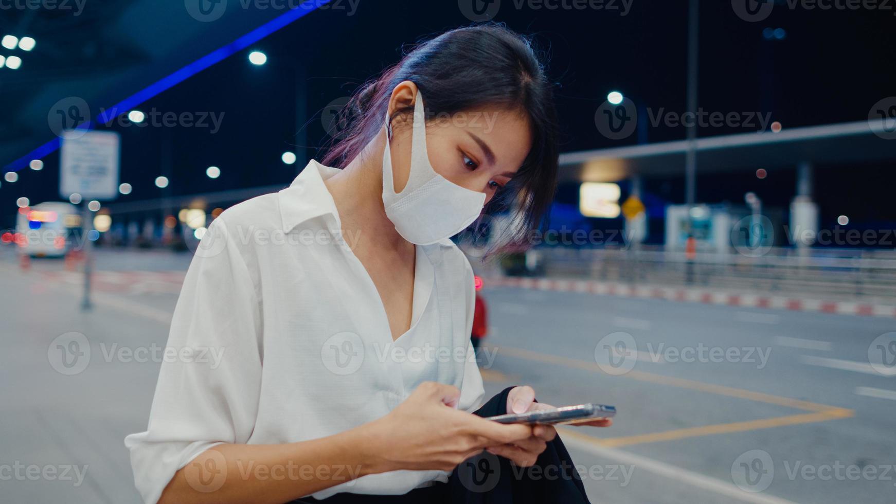 asiatisk affärsflicka anländer destination bär ansiktsmask stå utanför titta smart telefon vänta bilterminal på inrikes flygplats. affärspendlare covid -pandemi, affärsresor social distanseringskoncept. foto