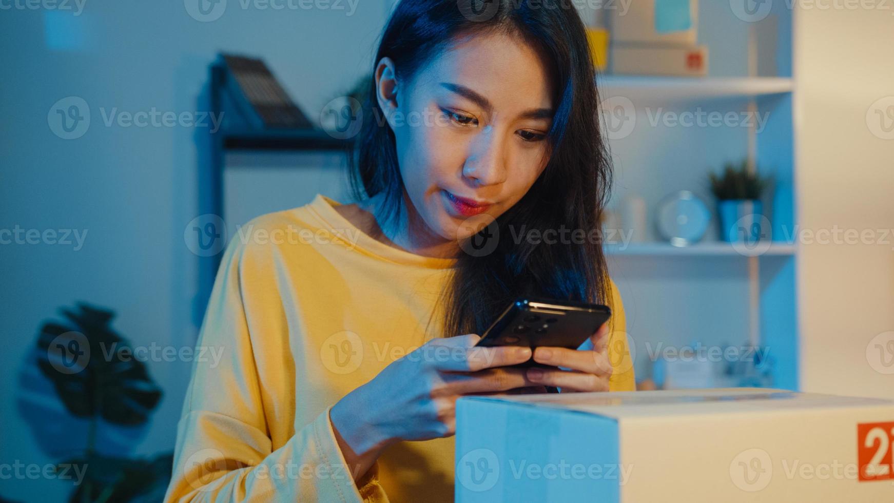 ung asiatisk kvinna använder smartphone ta streckkodsbild på paketprodukt för leverans till kund på hemmakontor på natten. småföretag, online marknadsföring, livsstil frilans koncept. foto