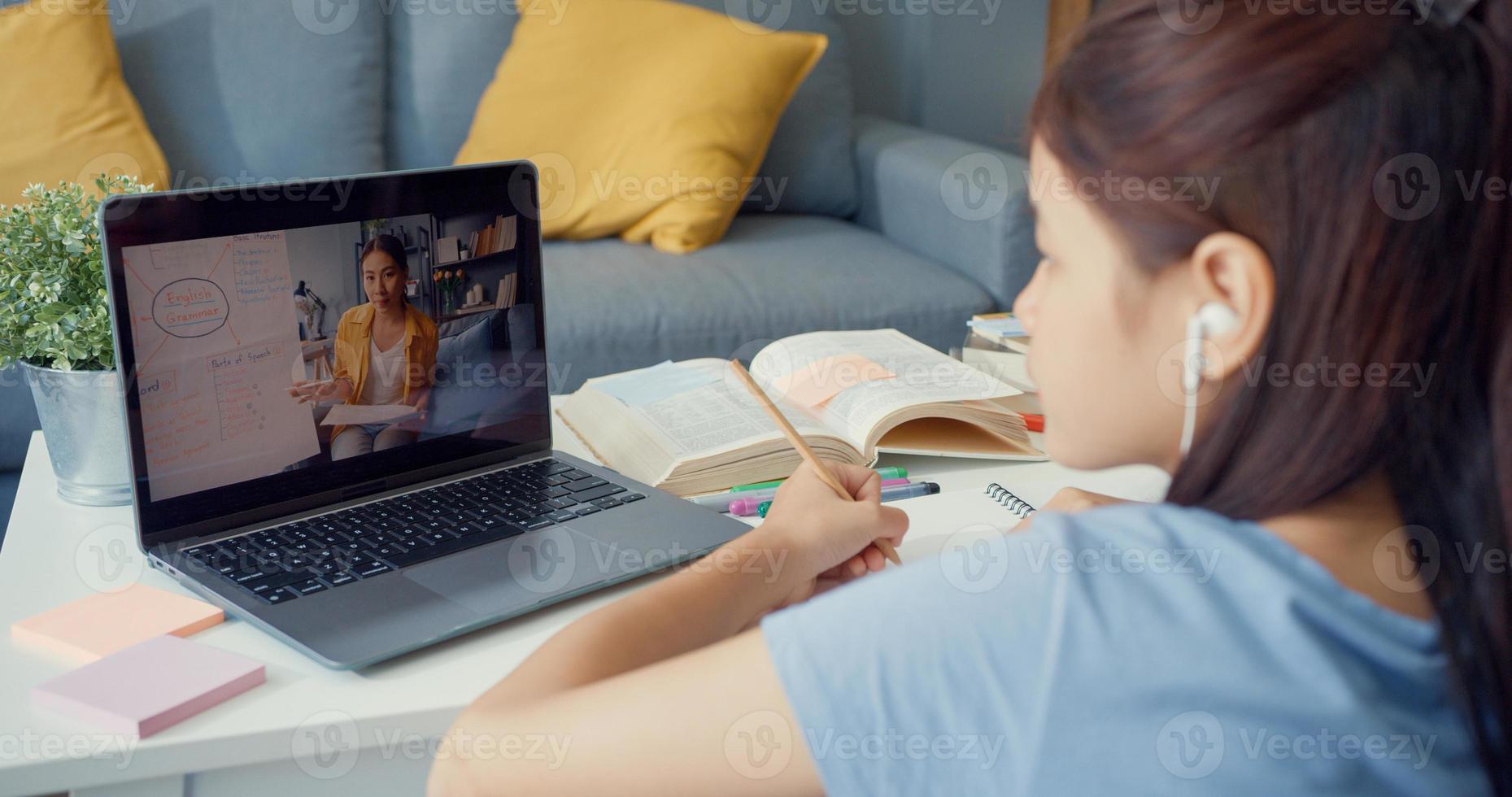 ung asiatisk tjej med tillfällig användning dator bärbar videosamtal lära online med lärare skriva föreläsning anteckningsbok vardagsrum hemma. isolera utbildning online e-learning coronavirus-pandemikoncept. foto