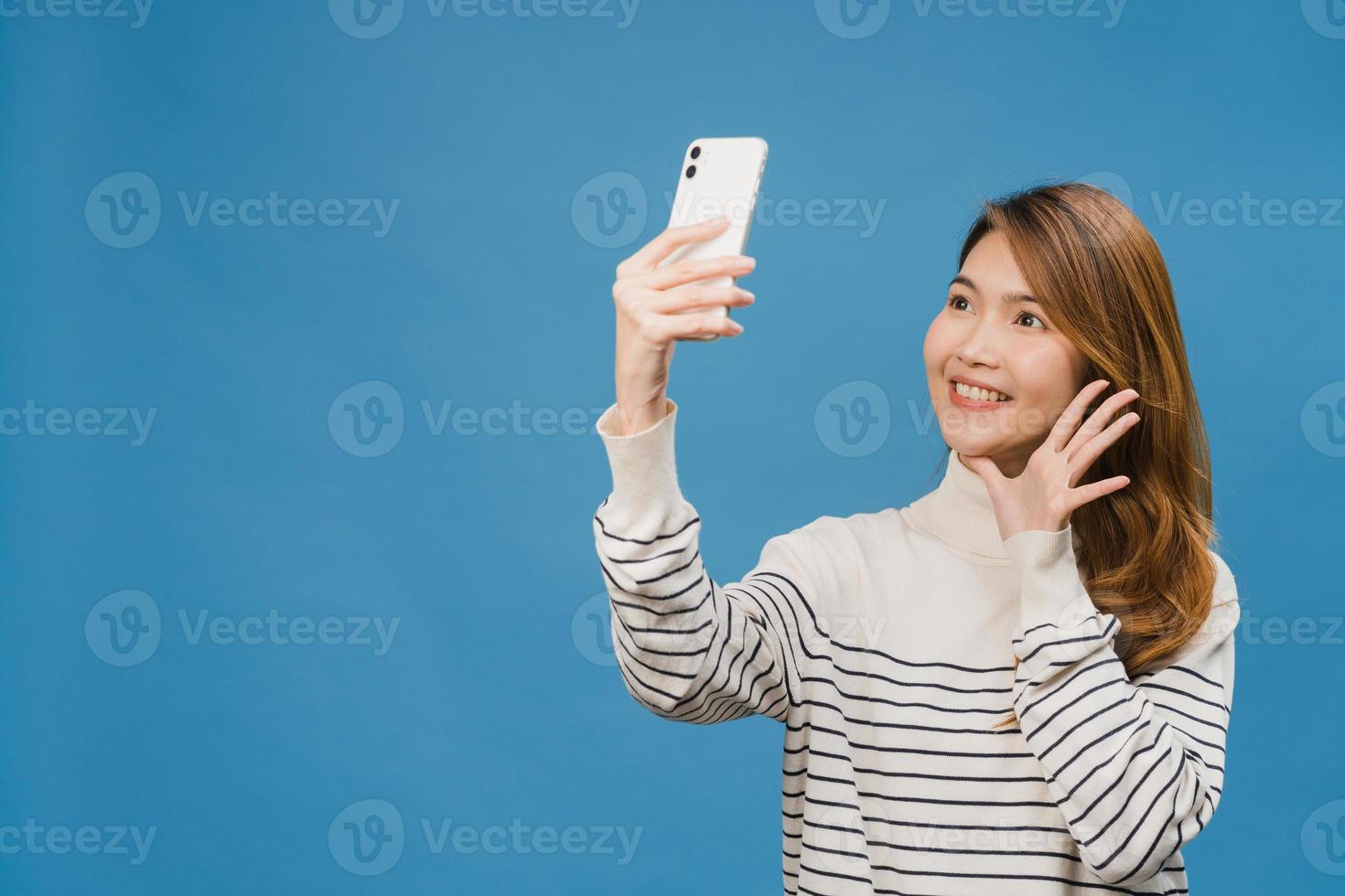 leende bedårande asiatisk kvinna som gör selfie -foto på smart telefon med positivt uttryck i vardagskläder och stativ isolerad på blå bakgrund. glad förtjusande glad kvinna jublar över framgång. foto