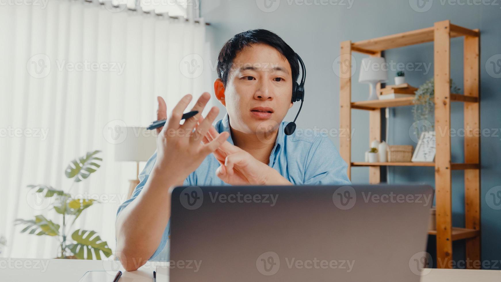 ung asiatisk affärsman bär hörlurar med bärbar dator prata med kollegor om planering i videosamtal medan du arbetar hemifrån i vardagsrummet. självisolering, social distansering, karantän för förebyggande av covid. foto