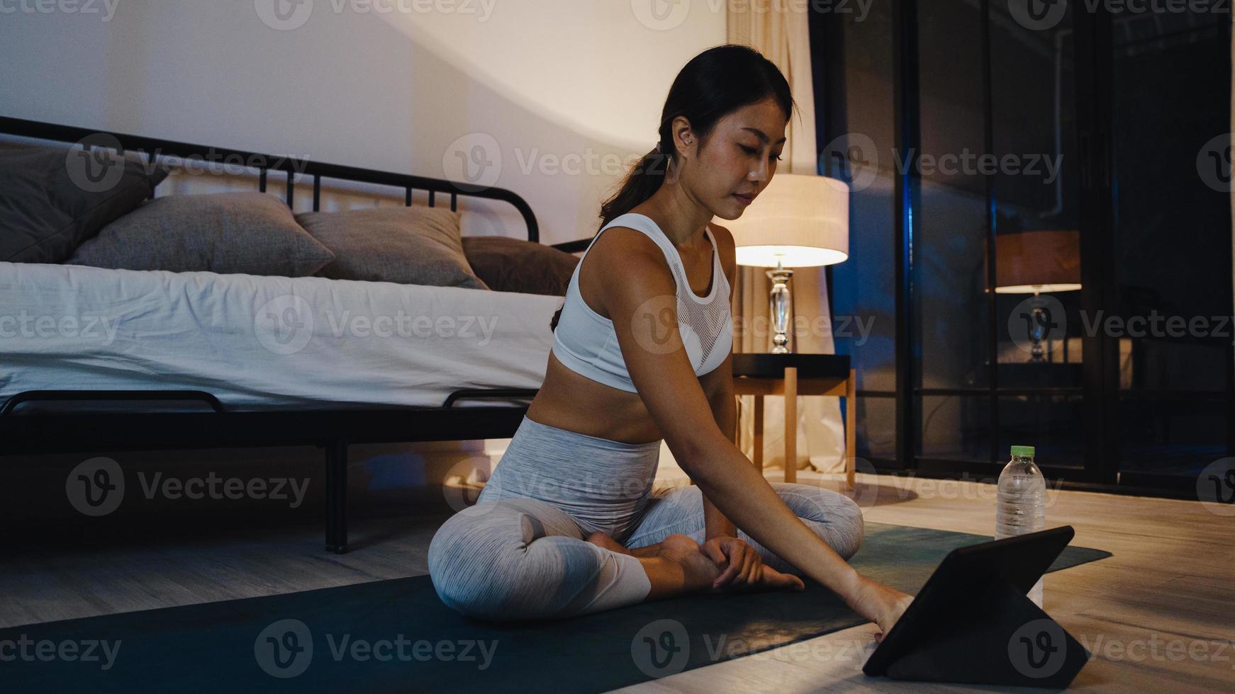 ung asiatisk dam i sportkläder övningar som tränar och använder surfplatta för att titta på yoga video tutorial hemma kväll. avlägsen utbildning med personlig tränare, social distans, online utbildningskoncept. foto