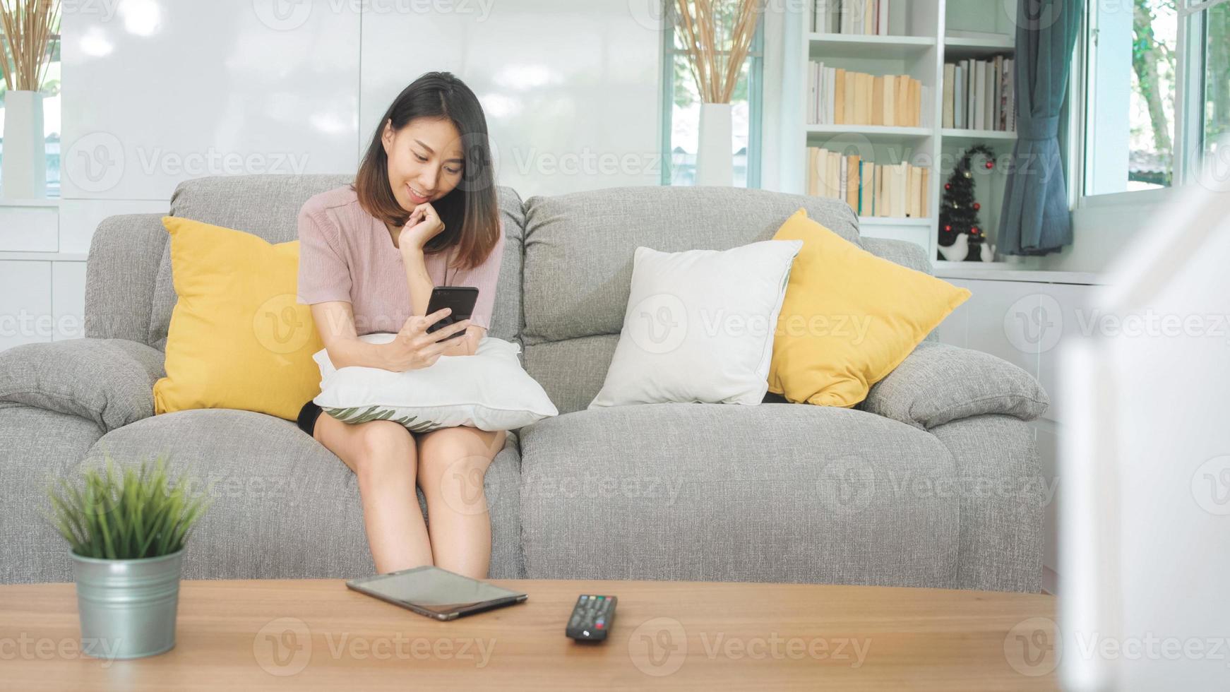 ung asiatisk kvinna som använder smartphone som kontrollerar sociala medier som känner sig lycklig leende medan hon ligger i soffan när hon kopplar av i vardagsrummet hemma. livsstil latin och spansktalande etnicitet kvinnor hemma koncept. foto