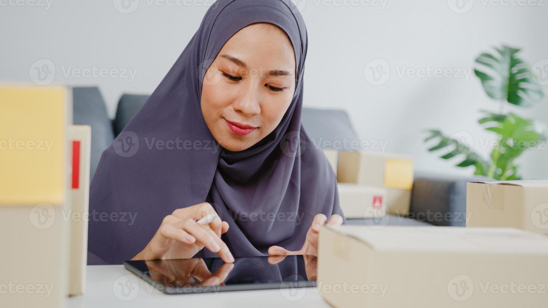 ung asien muslimsk affärskvinna kontrollera produkt inköpsorder på lager och spara till surfplatta arbete på hemmakontor. småföretagare, onlinemarknadsleverans, livsstil frilansande koncept. foto