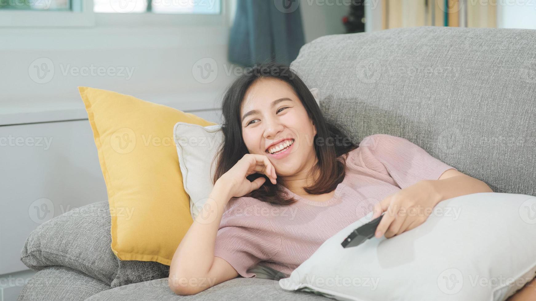 ung asiatisk tonårskvinna som tittar på tv hemma, kvinnlig känner sig lycklig liggande på soffan i vardagsrummet. livsstil kvinna koppla av på morgonen hemma koncept. foto