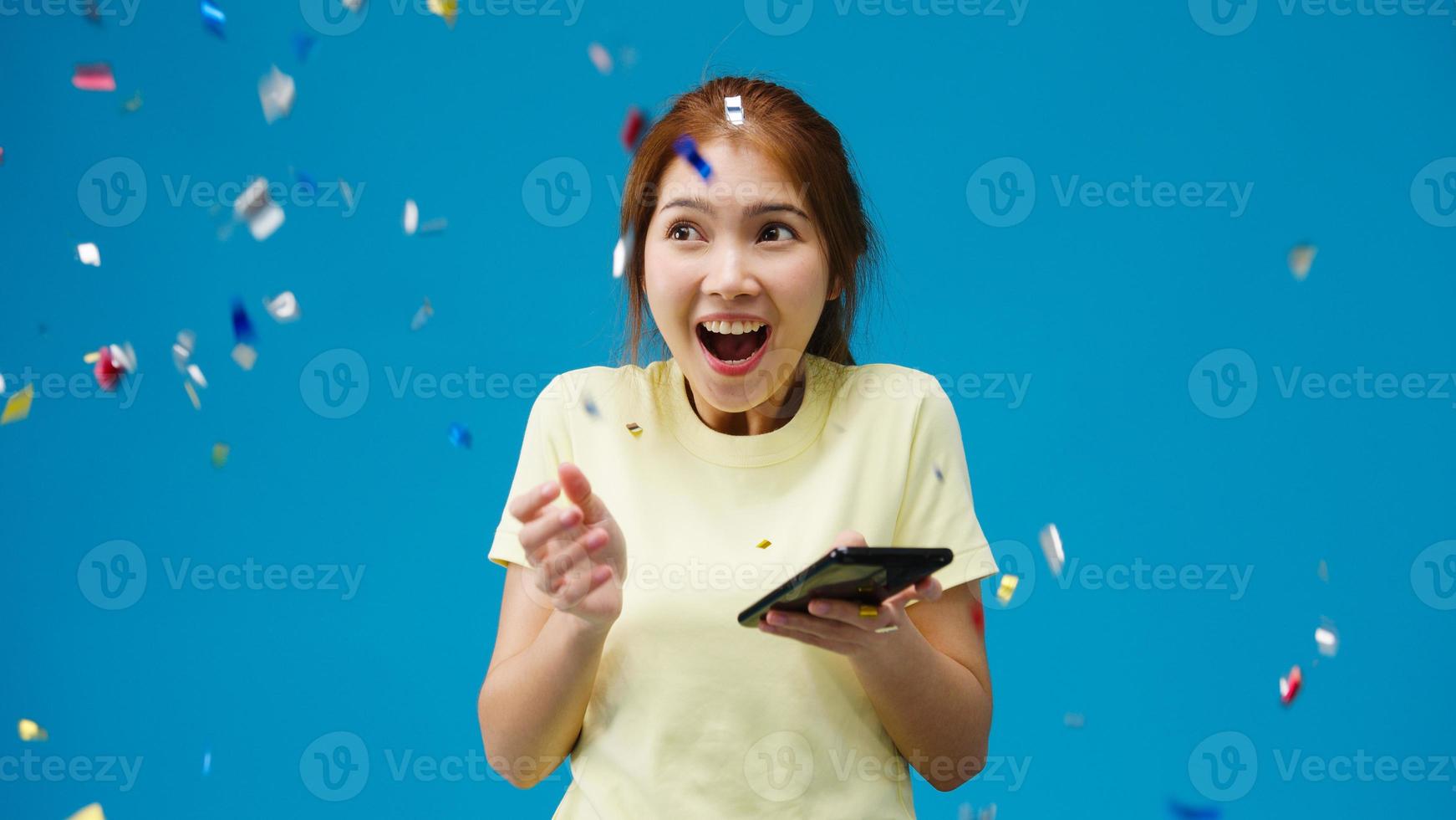 förvånad ung asiatisk dam som använder mobiltelefon med positivt uttryck, ler brett, klädd i avslappnad trasa under konfettiregn och firar på blå bakgrund. glad glad kvinna jublar över framgång. foto