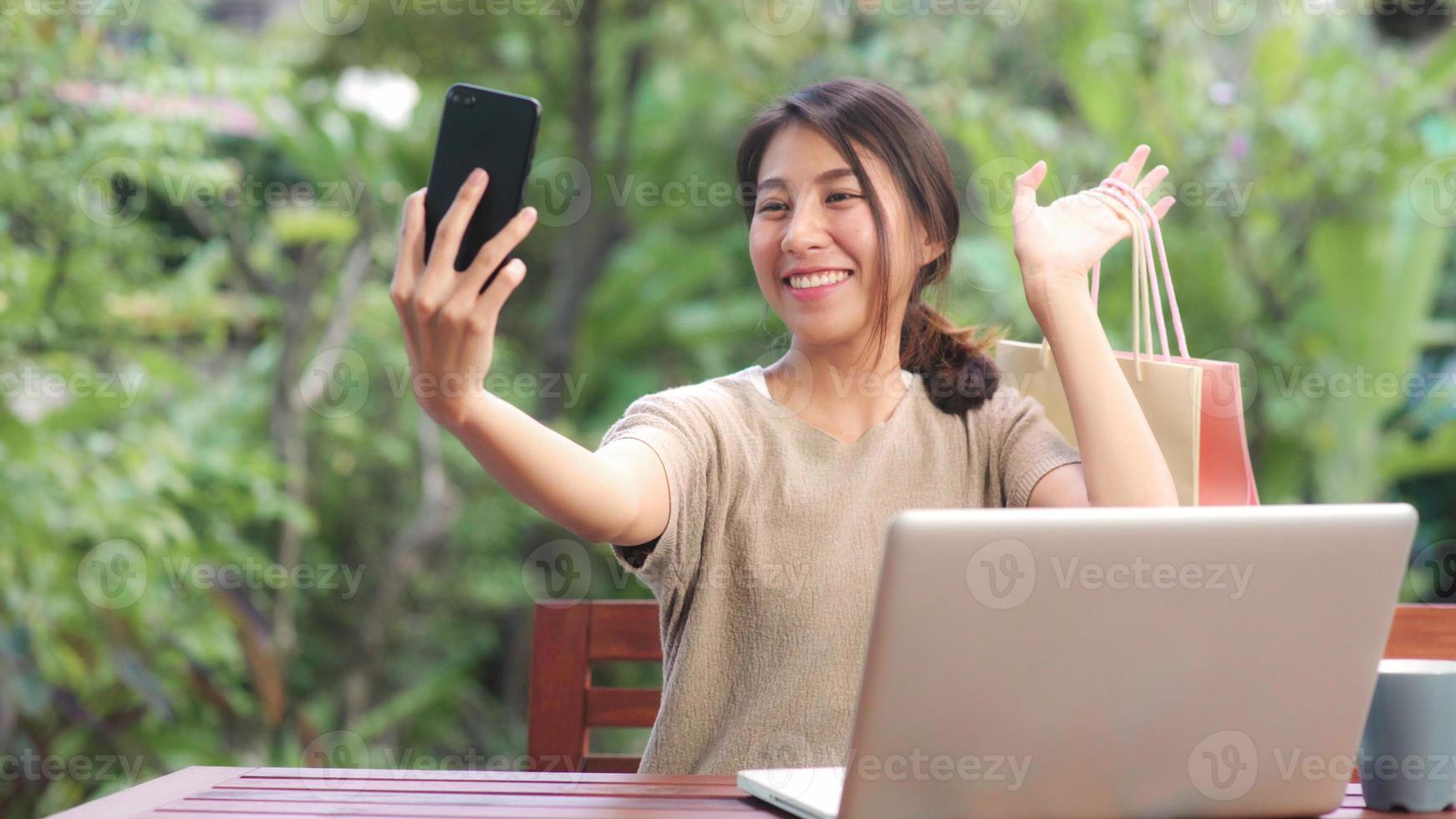 asiatisk kvinna som använder mobiltelefon selfie inlägg i sociala medier, kvinnlig koppla av känner sig glad och visar shoppingkassar som sitter på bordet i trädgården på morgonen. livsstil kvinnor slappna av hemma koncept. foto