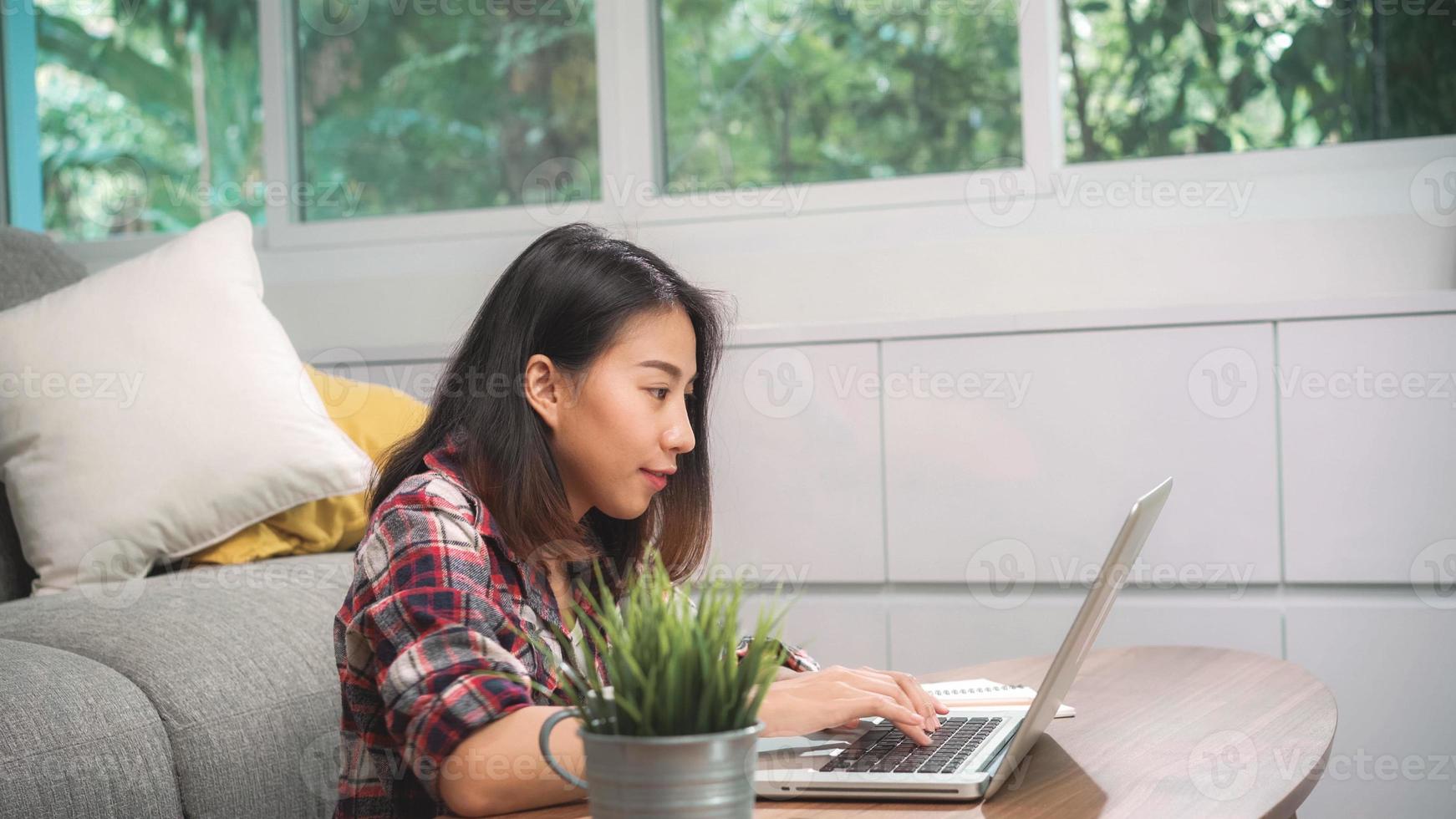 ung affärs frilans asiatisk kvinna som arbetar på bärbar dator som kontrollerar sociala medier medan hon ligger i soffan när hon kopplar av i vardagsrummet hemma. livsstil latin och spansktalande etnicitet kvinnor hemma koncept. foto