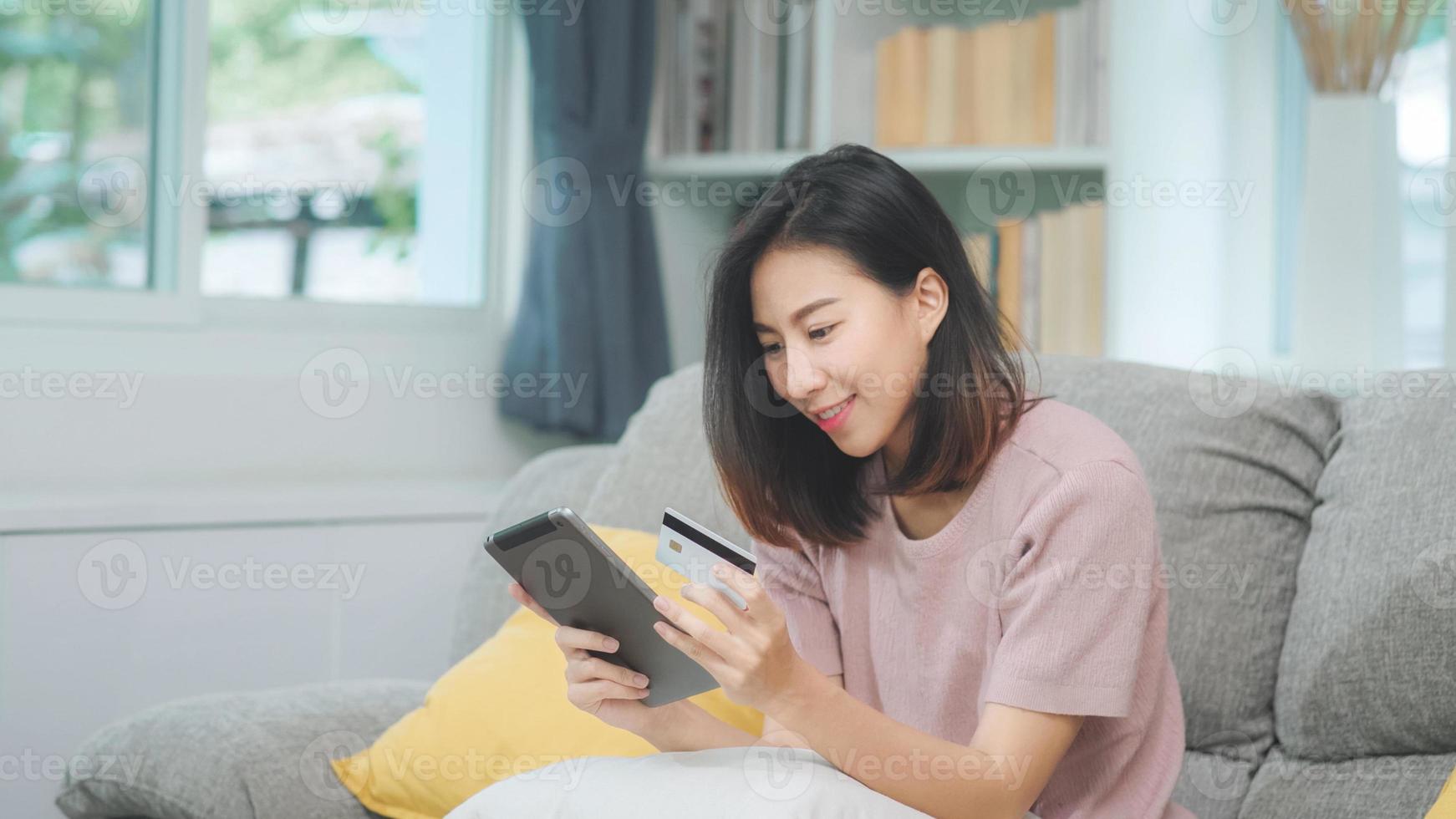 ung leende asiatisk kvinna som använder surfplatta och köper onlineshopping med kreditkort medan hon ligger på soffan när hon kopplar av i vardagsrummet hemma. livsstil latin och spansktalande etnicitet kvinnor hemma koncept. foto